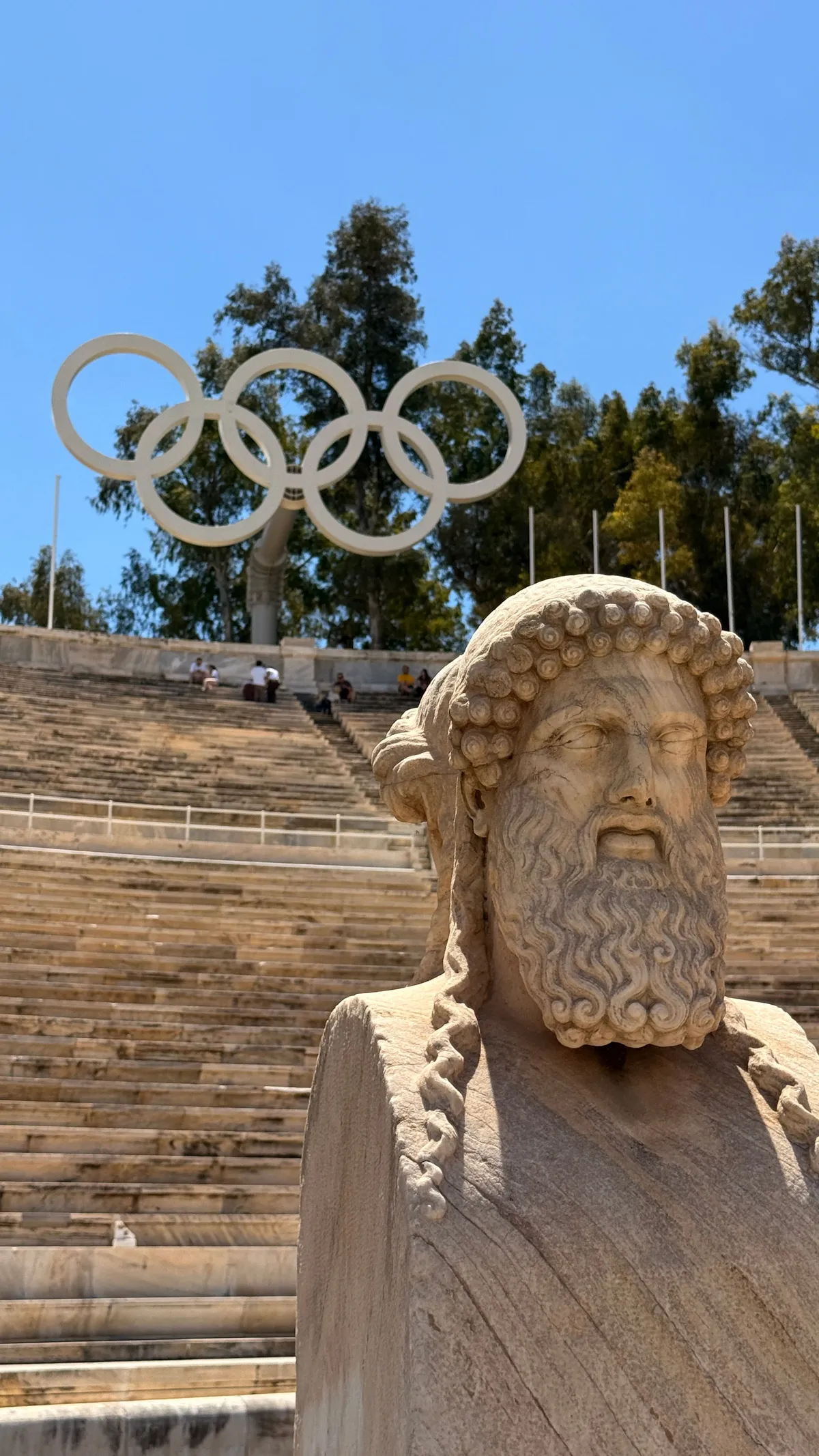 Marble bust of a bearded man at the Panathenaic Stadium with the Olympic rings on a pole behind