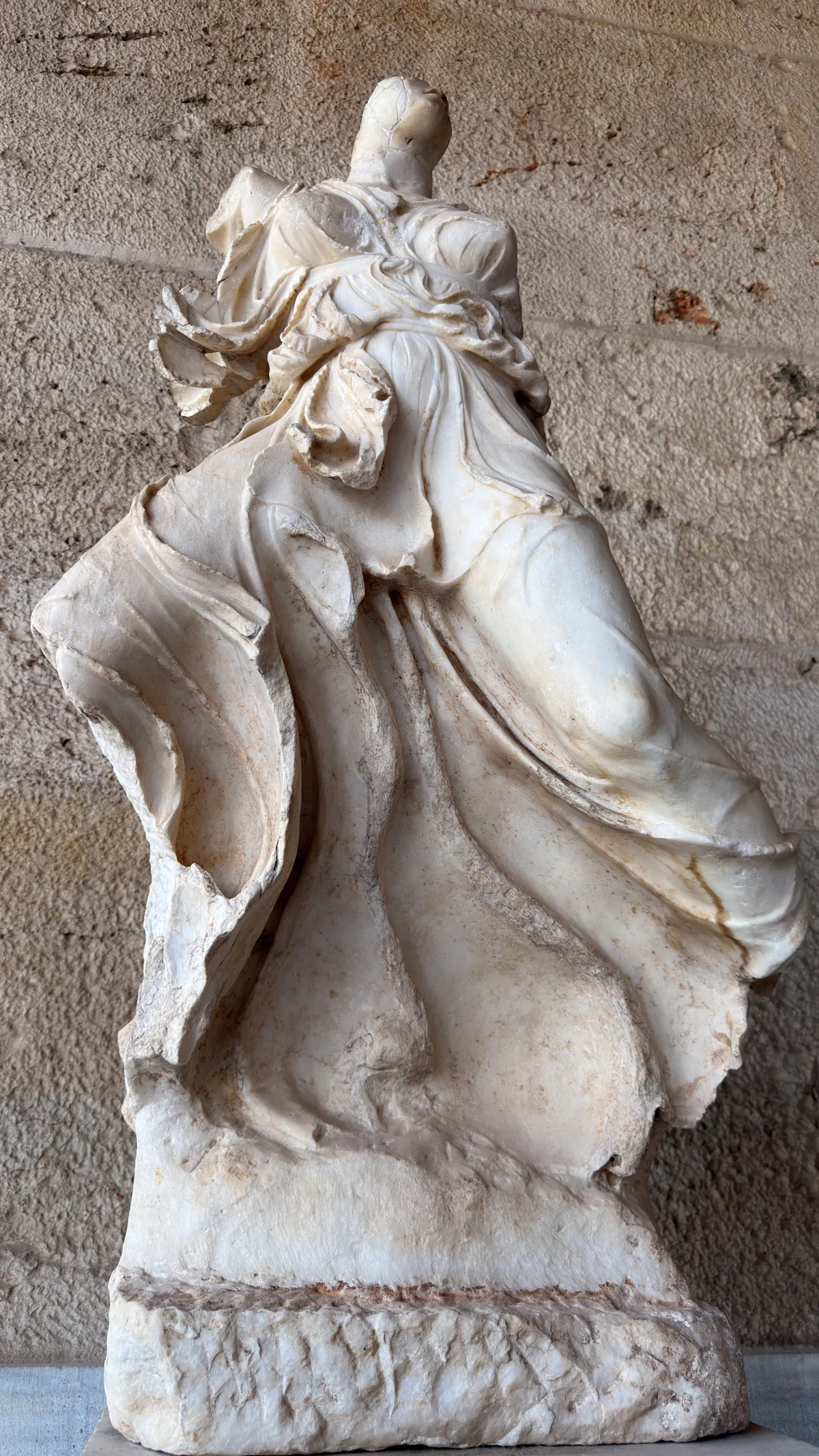 Headless marble statue with deeply folded drapery in front of a stone wall