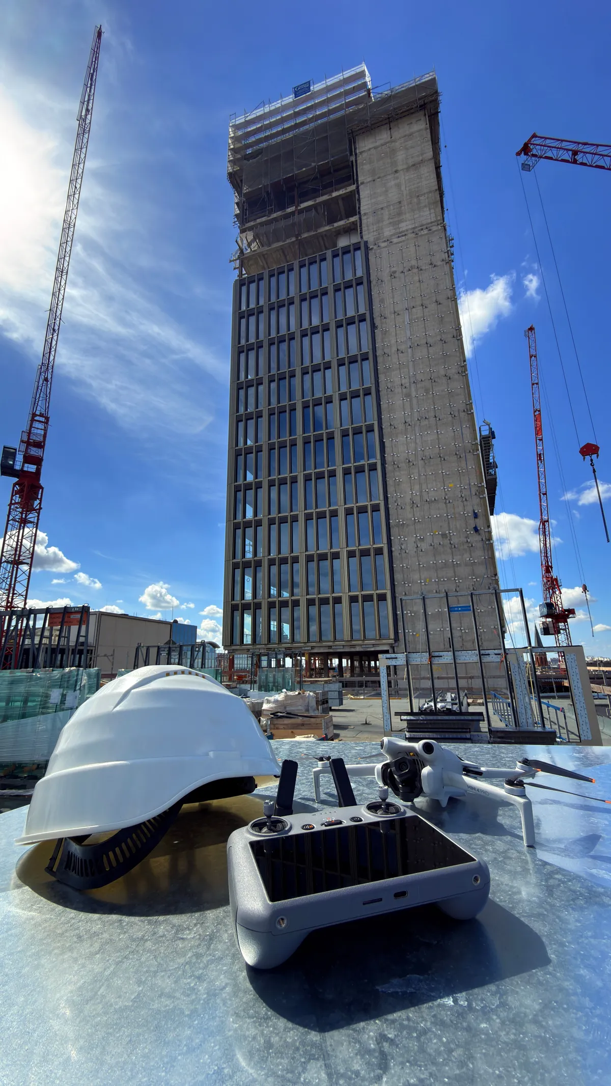 High-rise under construction with cranes against a blue sky, a white hard hat and drone controller on the ledge in front