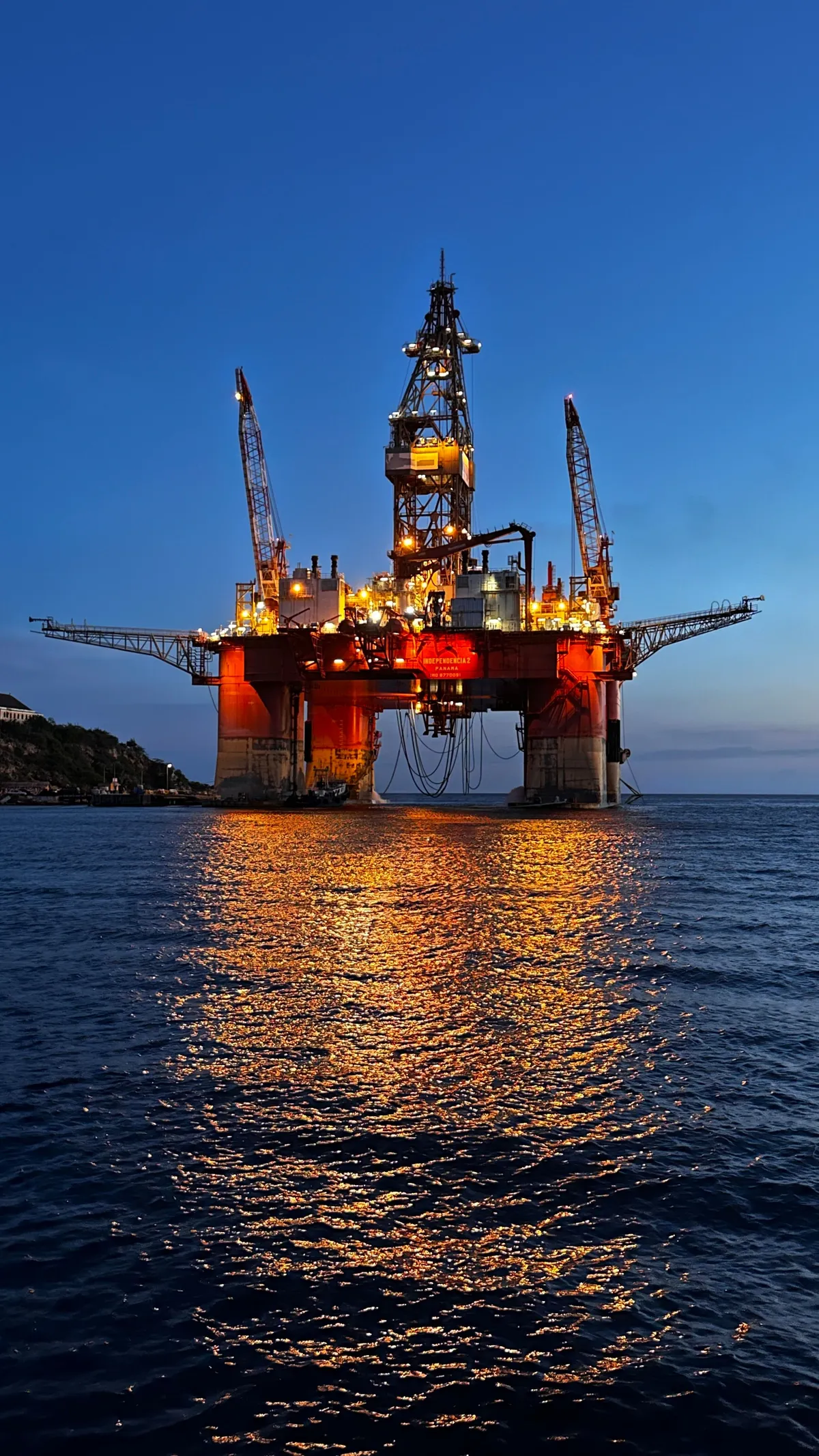 Illuminated oil rig at blue hour anchored off the coast, its lights reflected in the sea