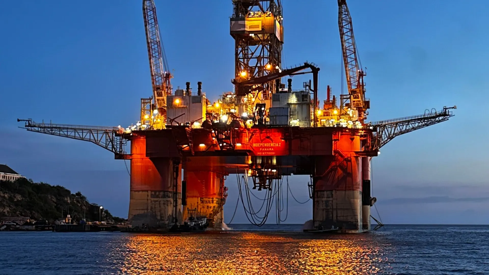 Illuminated oil rig at blue hour anchored off the coast, its lights reflected in the sea