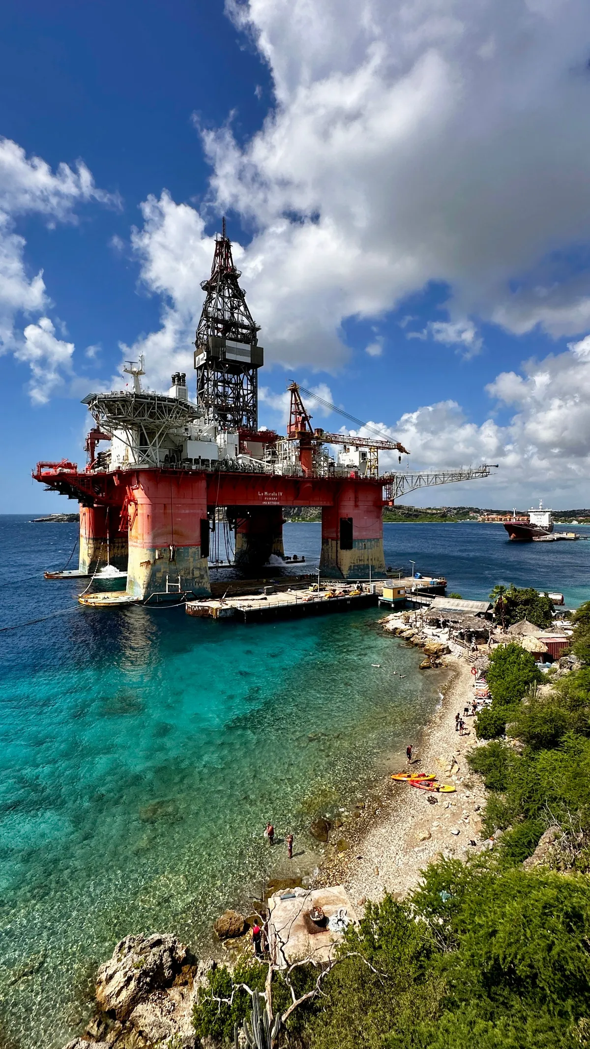 Retired oil rig above turquoise water and a rocky shoreline under cumulus clouds