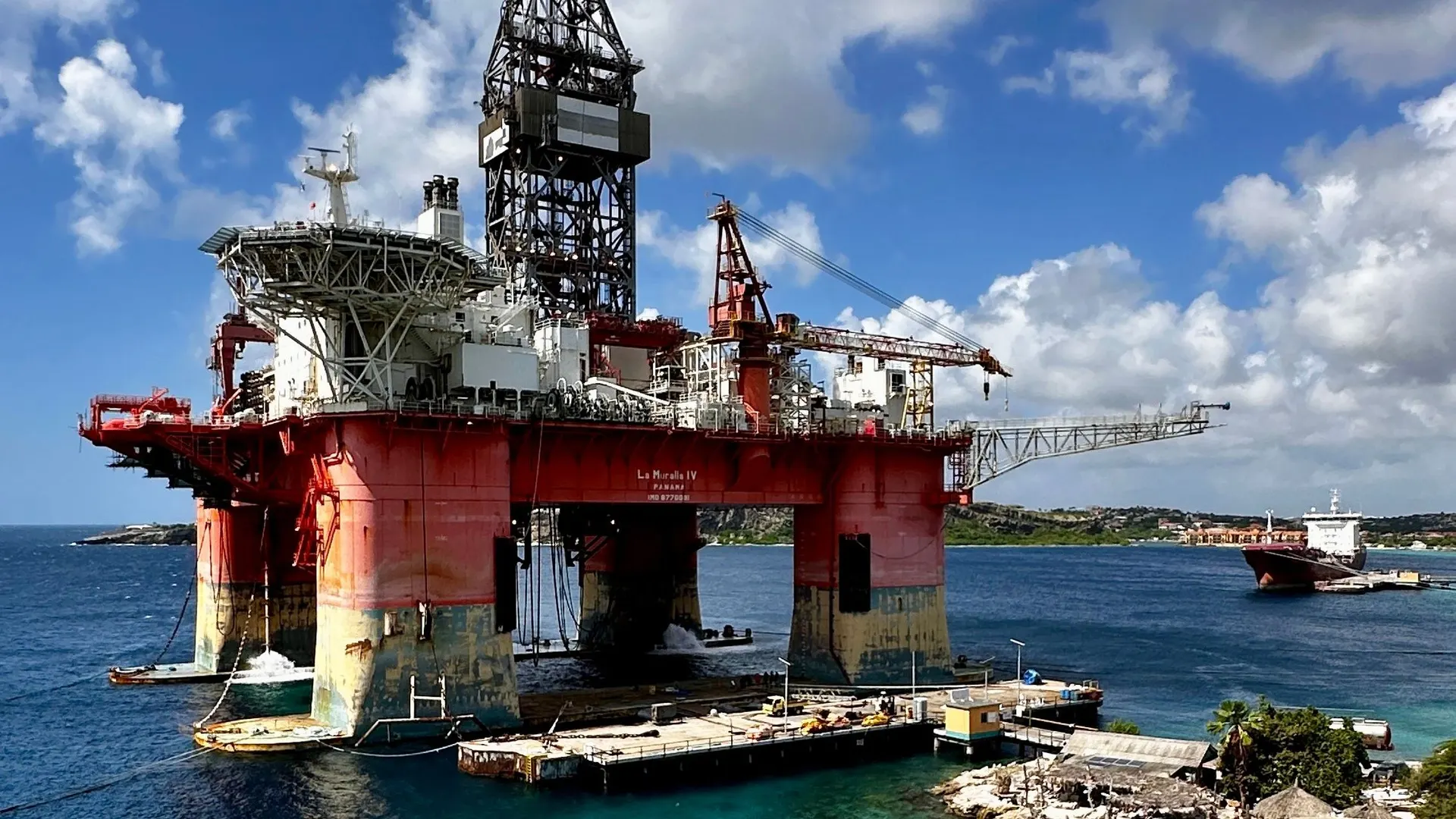 Retired oil rig above turquoise water and a rocky shoreline under cumulus clouds