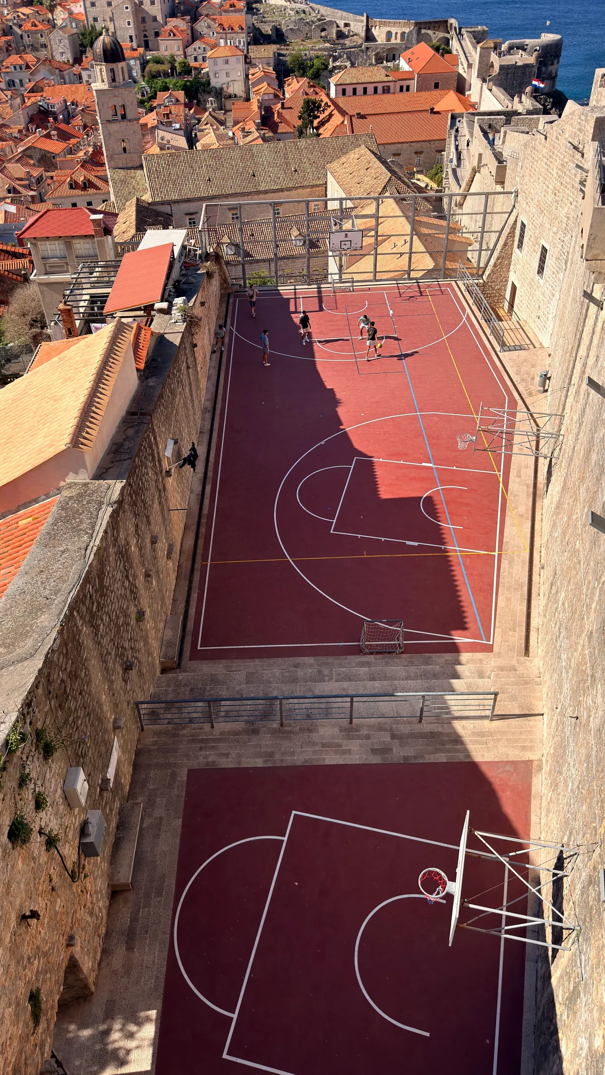 Red basketball courts wedged between Dubrovnik old-town walls and terracotta roofs, seen from the city wall