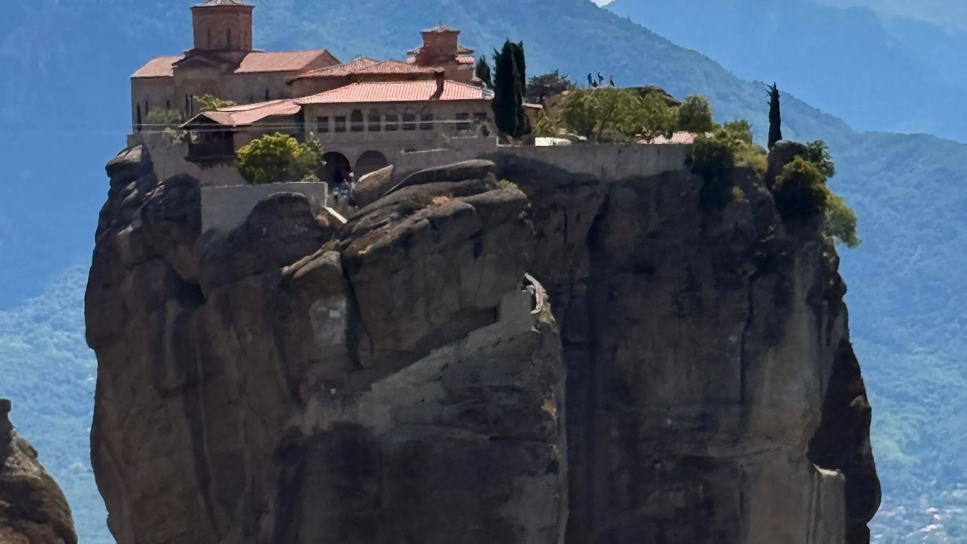 Meteora monastery of the Holy Trinity on a free-standing rock pillar above a green valley, mountains behind
