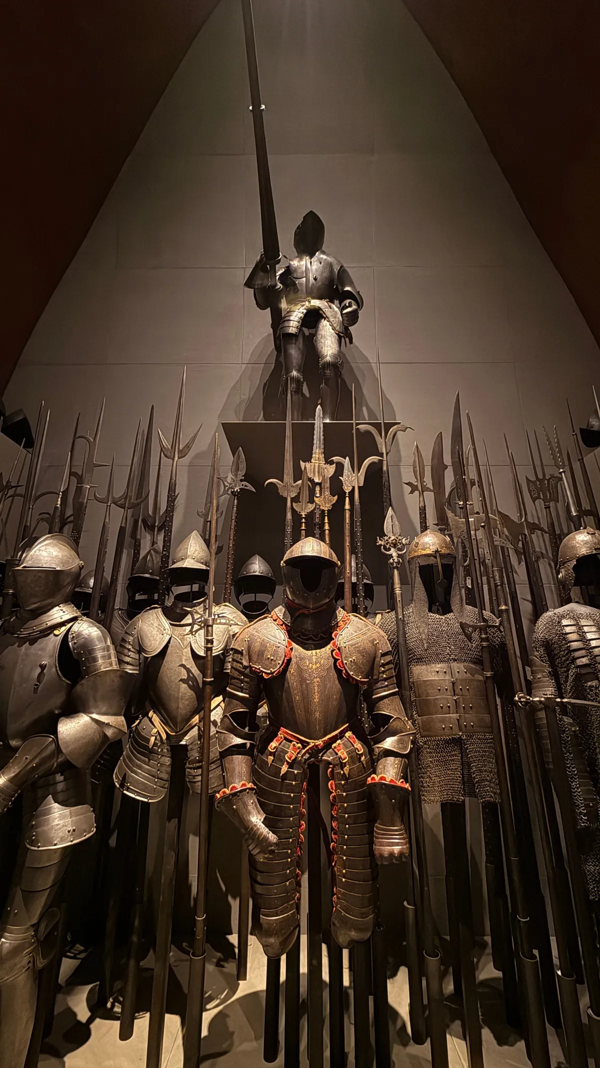 Suits of armour and polearms densely staged in a dark museum hall, one knight lit from above