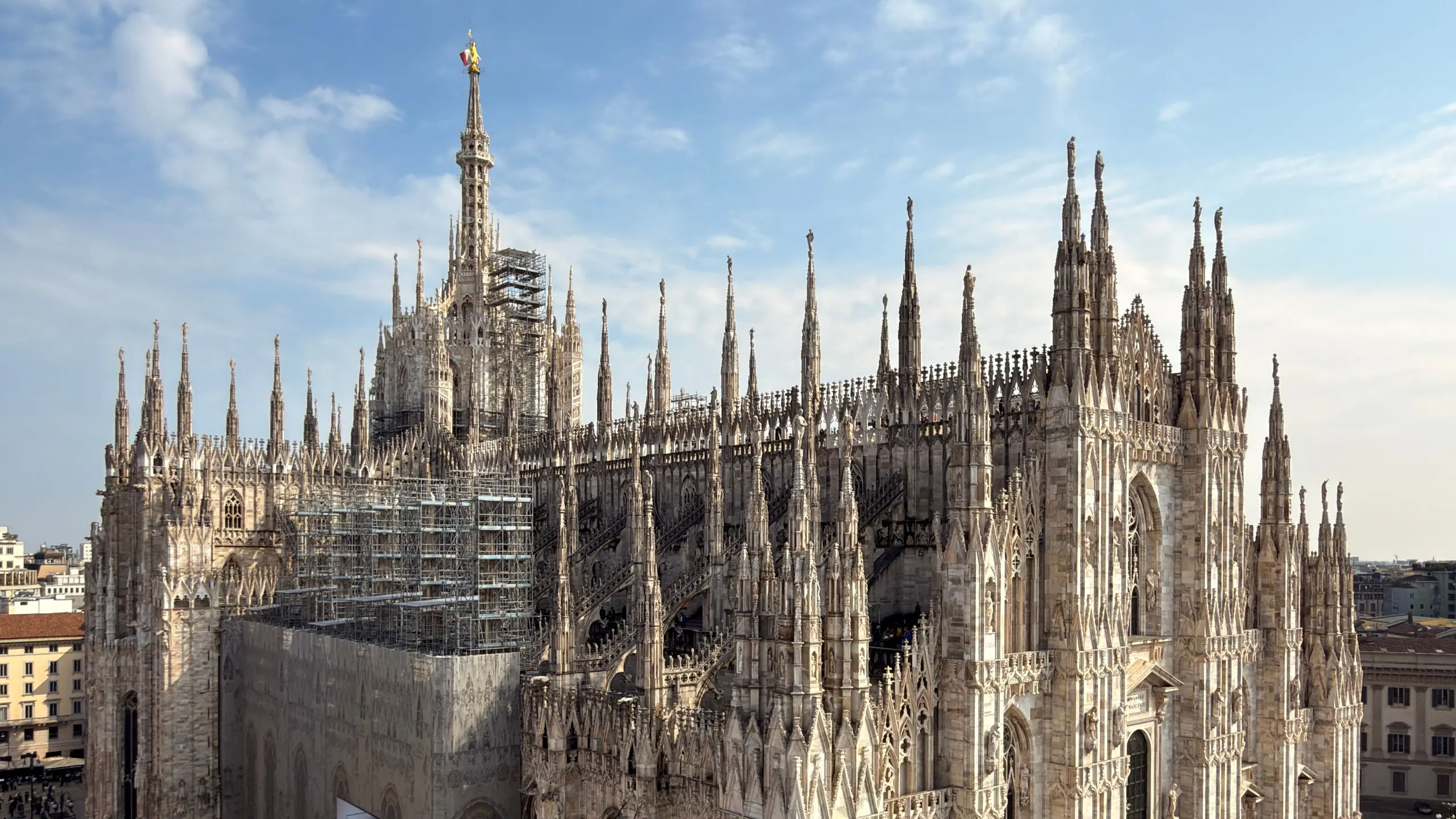 Milan Cathedral seen from a rooftop terrace, pinnacles against the sky, the square with people below