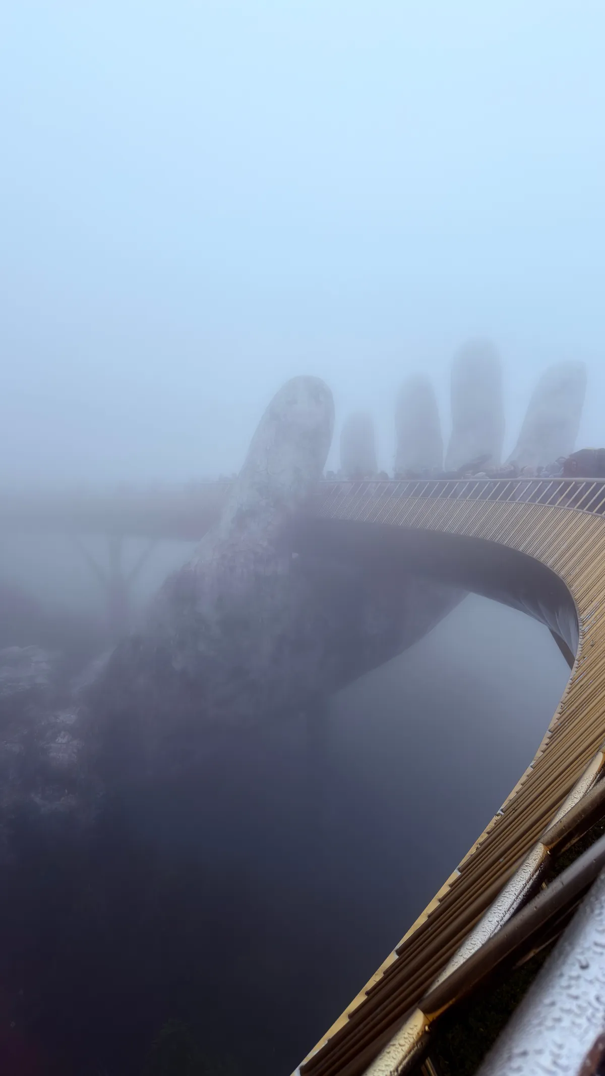 The Golden Bridge at Ba Na Hills disappearing into dense fog, a giant stone hand holding the walkway