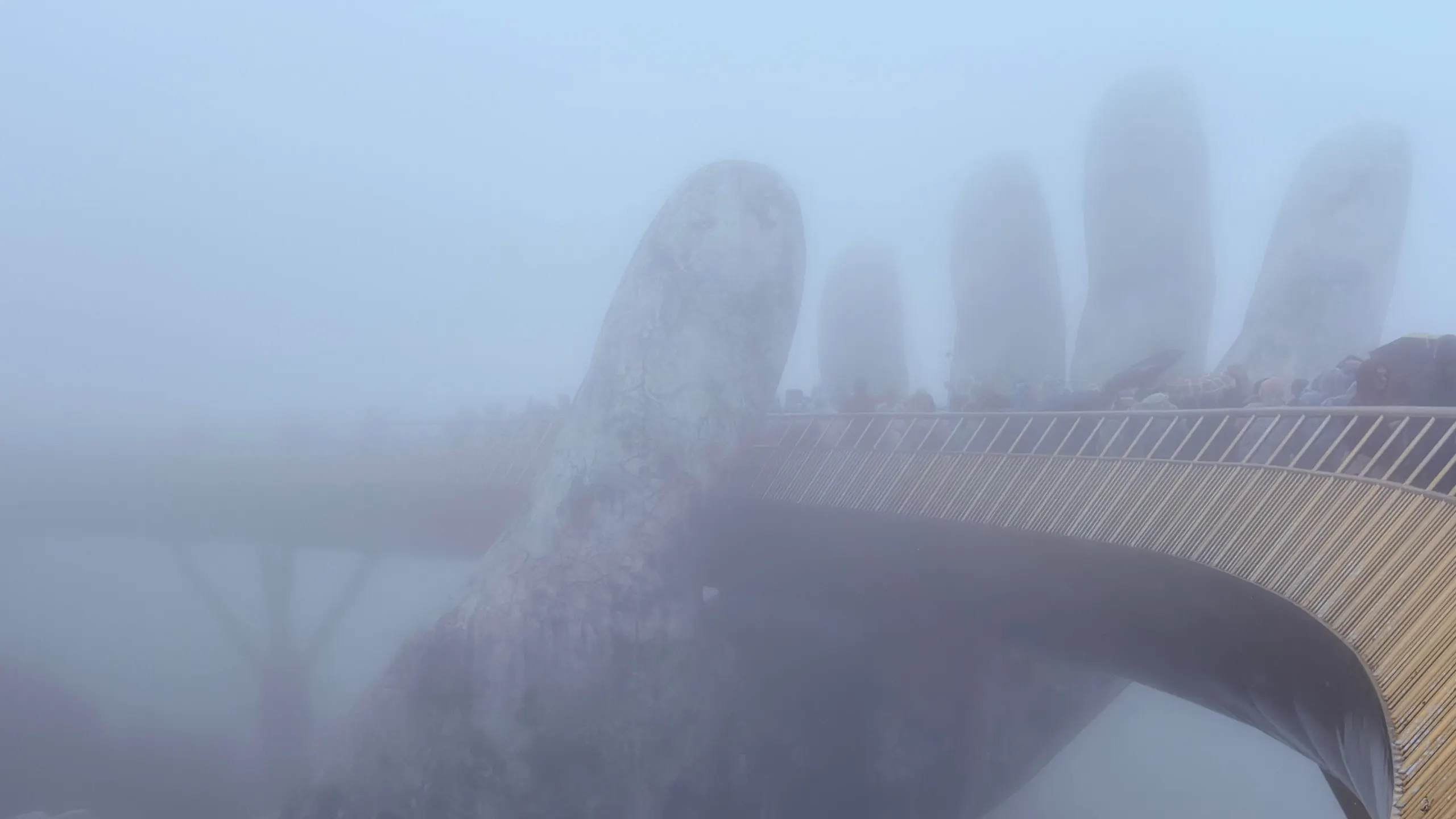 The Golden Bridge at Ba Na Hills disappearing into dense fog, a giant stone hand holding the walkway