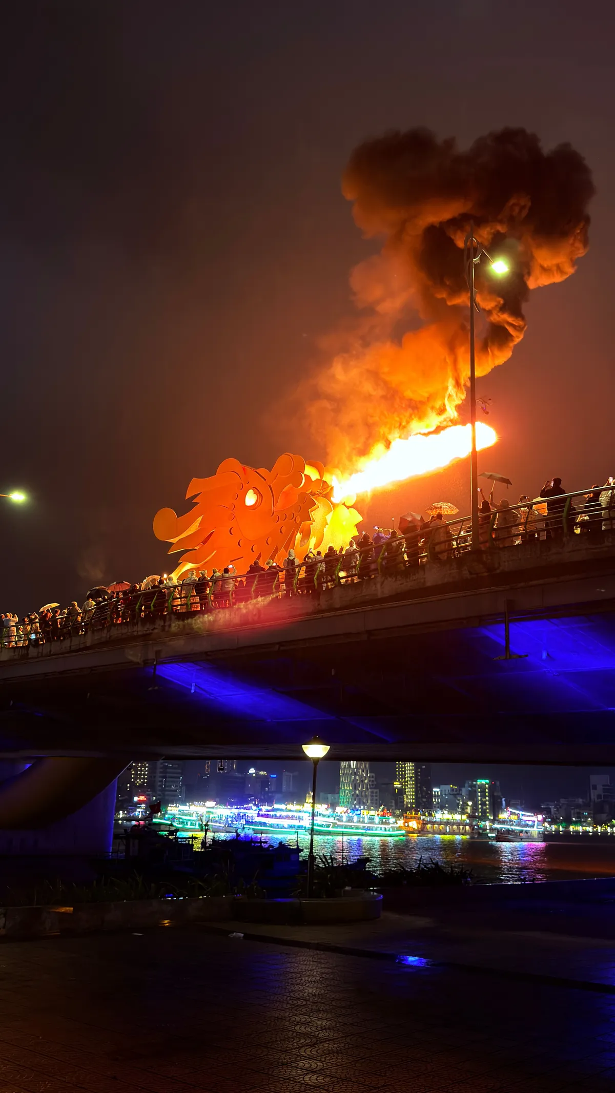 The Dragon Bridge in Da Nang spitting fire at night, a crowd watching from the promenade, the river lit in blue