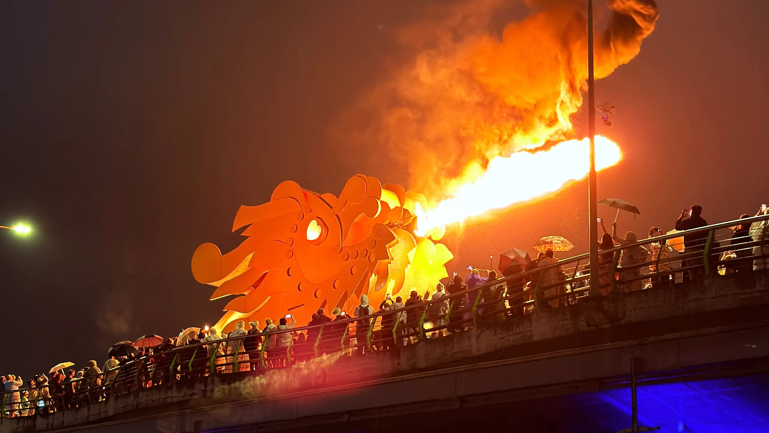 The Dragon Bridge in Da Nang spitting fire at night, a crowd watching from the promenade, the river lit in blue
