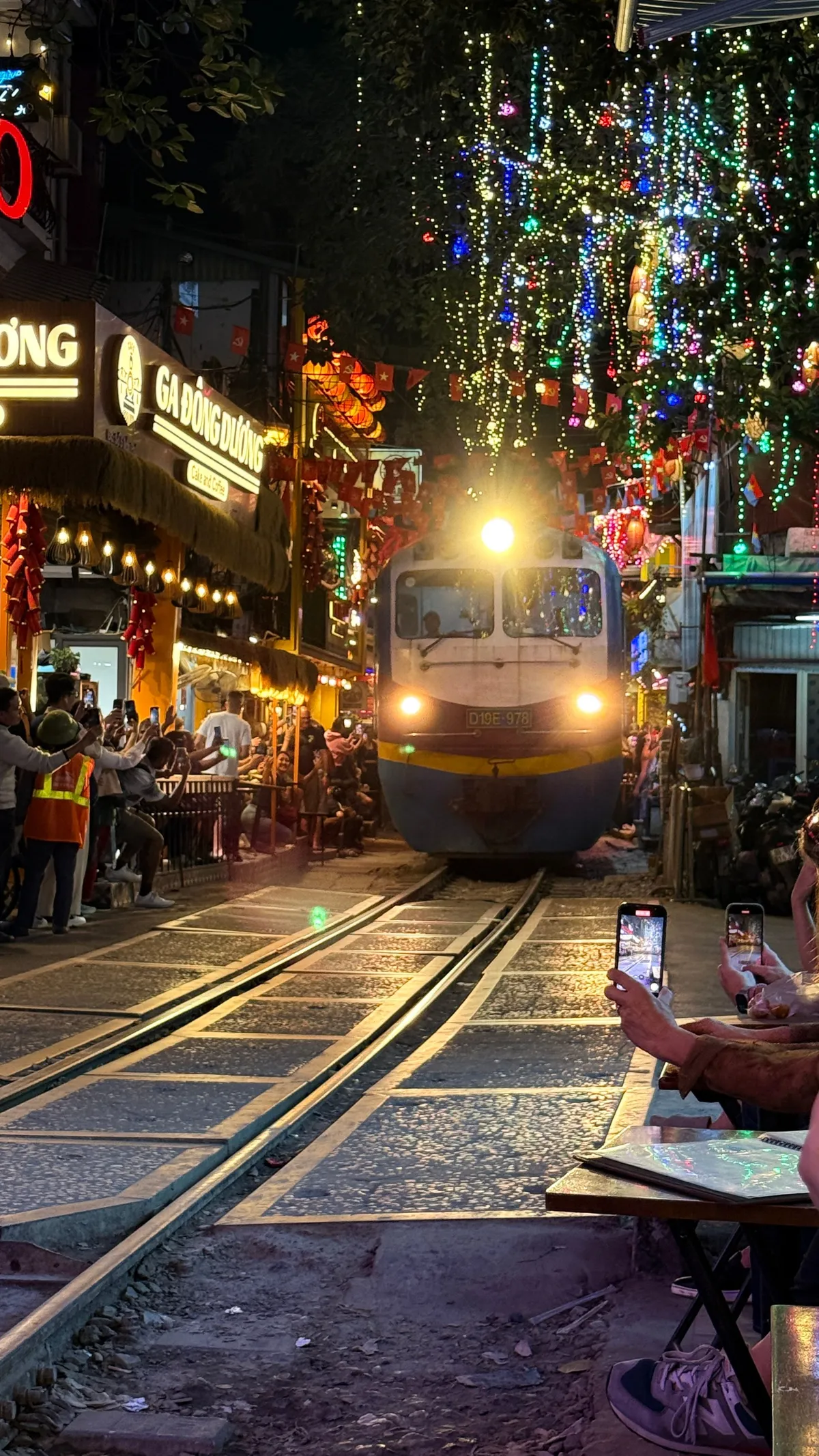 Train approaching along Hanoi’s train street at night, cafés lit up on both sides, people filming from the tracks