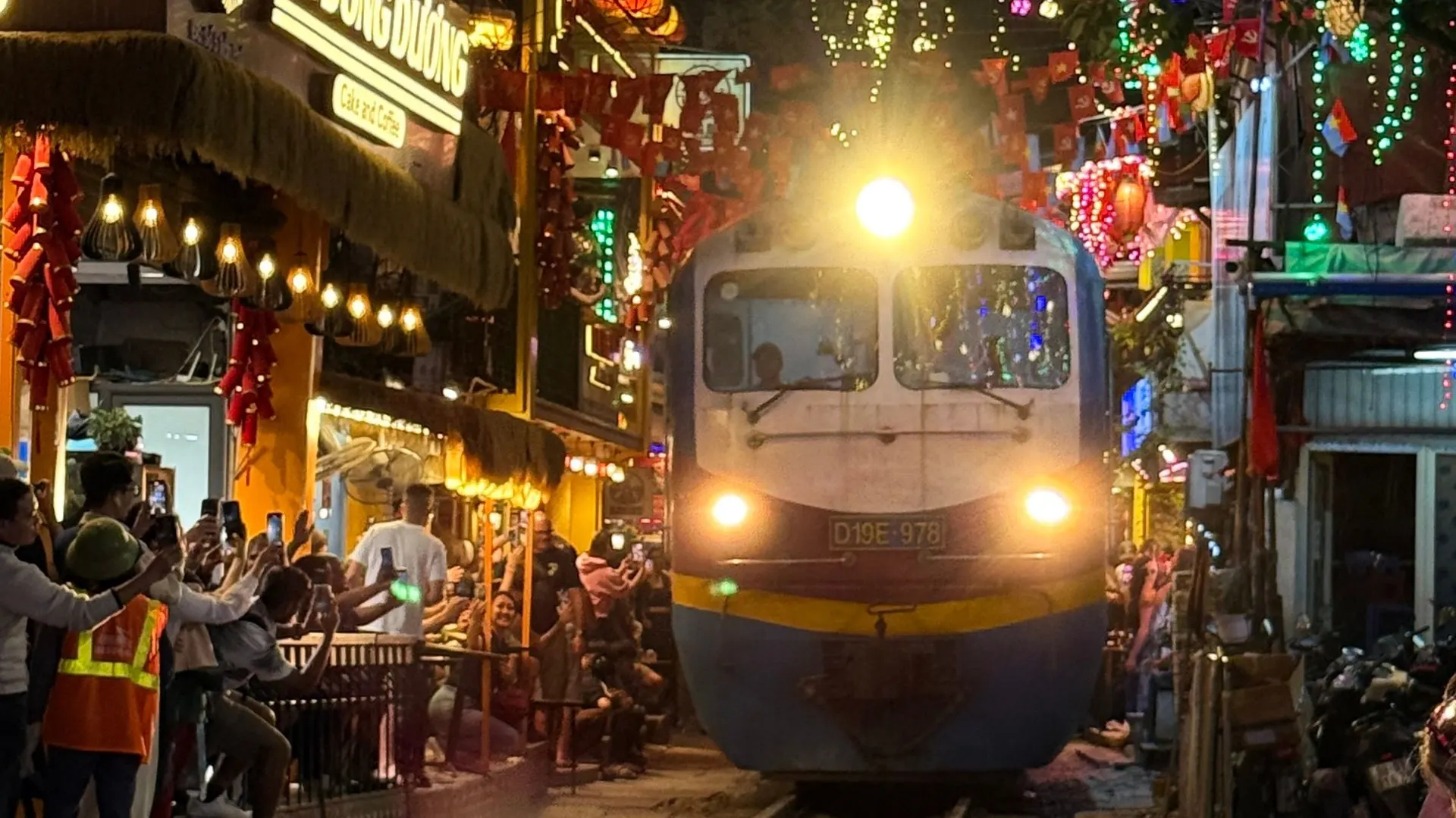 Train approaching along Hanoi’s train street at night, cafés lit up on both sides, people filming from the tracks