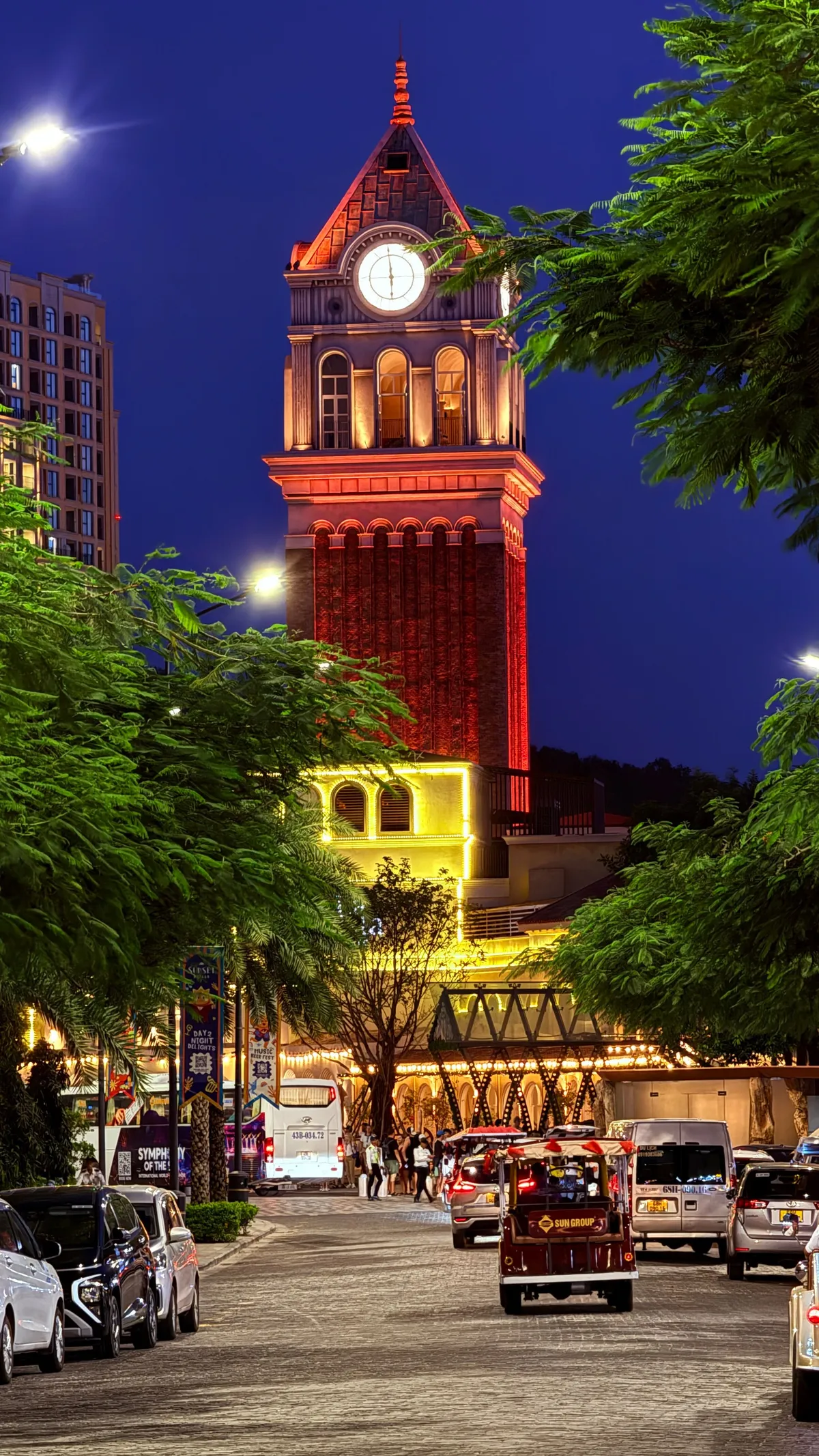Illuminated brick clock tower at Grand World Phu Quoc at blue hour, palm trees and tuk-tuks in the street