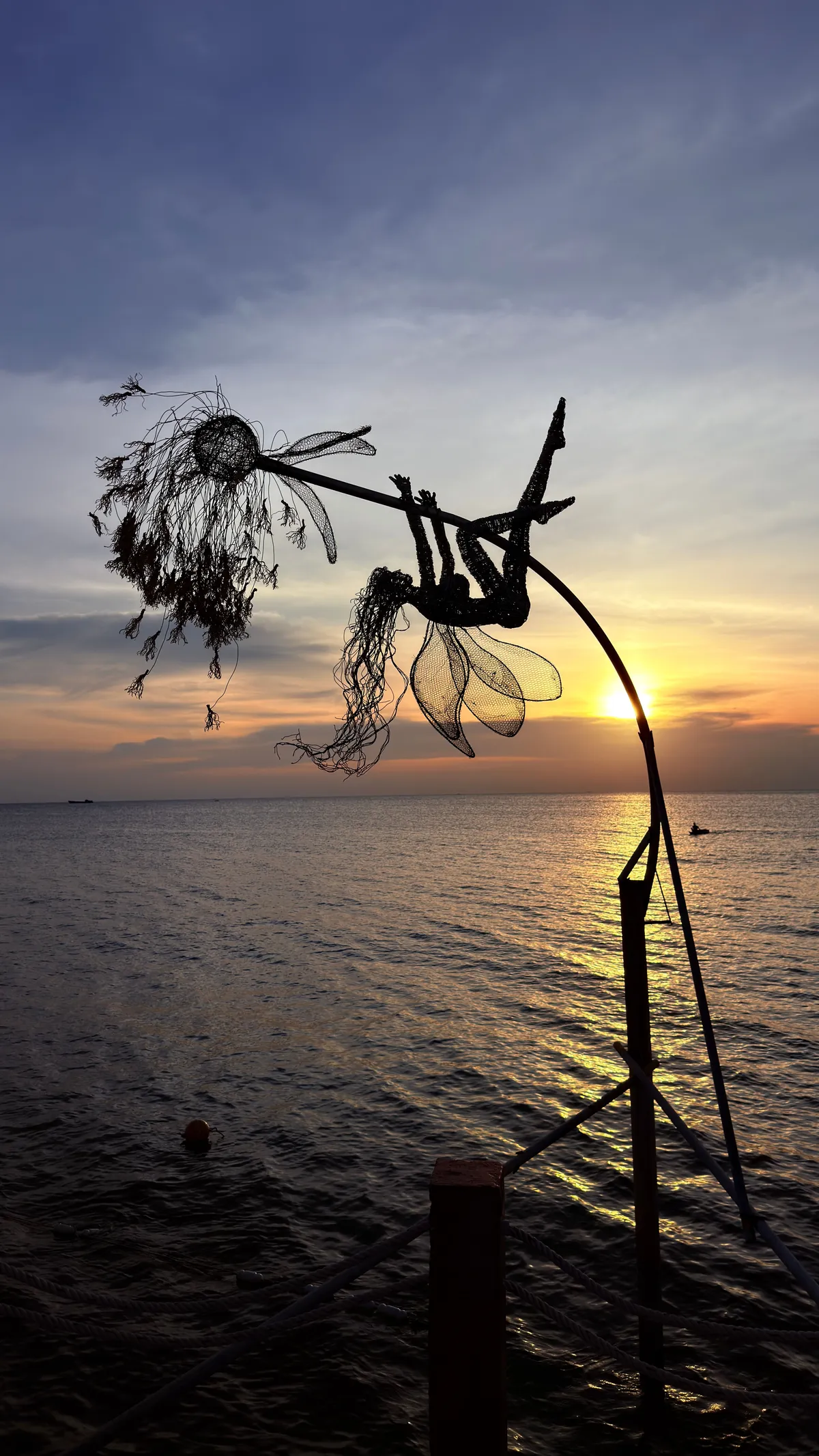 Wire sculpture of a fairy blowing a dandelion in silhouette against the evening sky over the sea