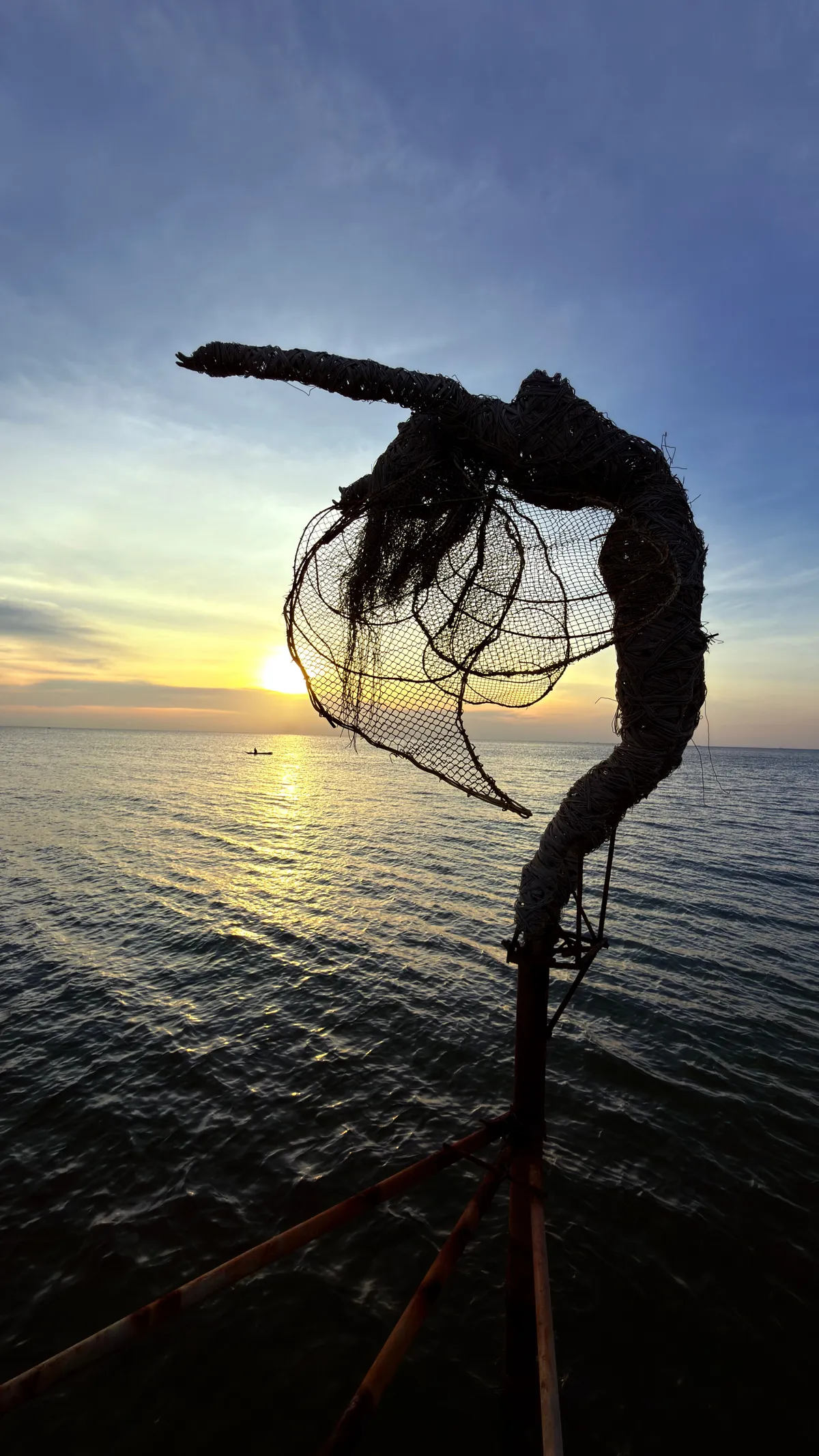 Wire sculpture of a figure holding a net in silhouette above the sea at dusk