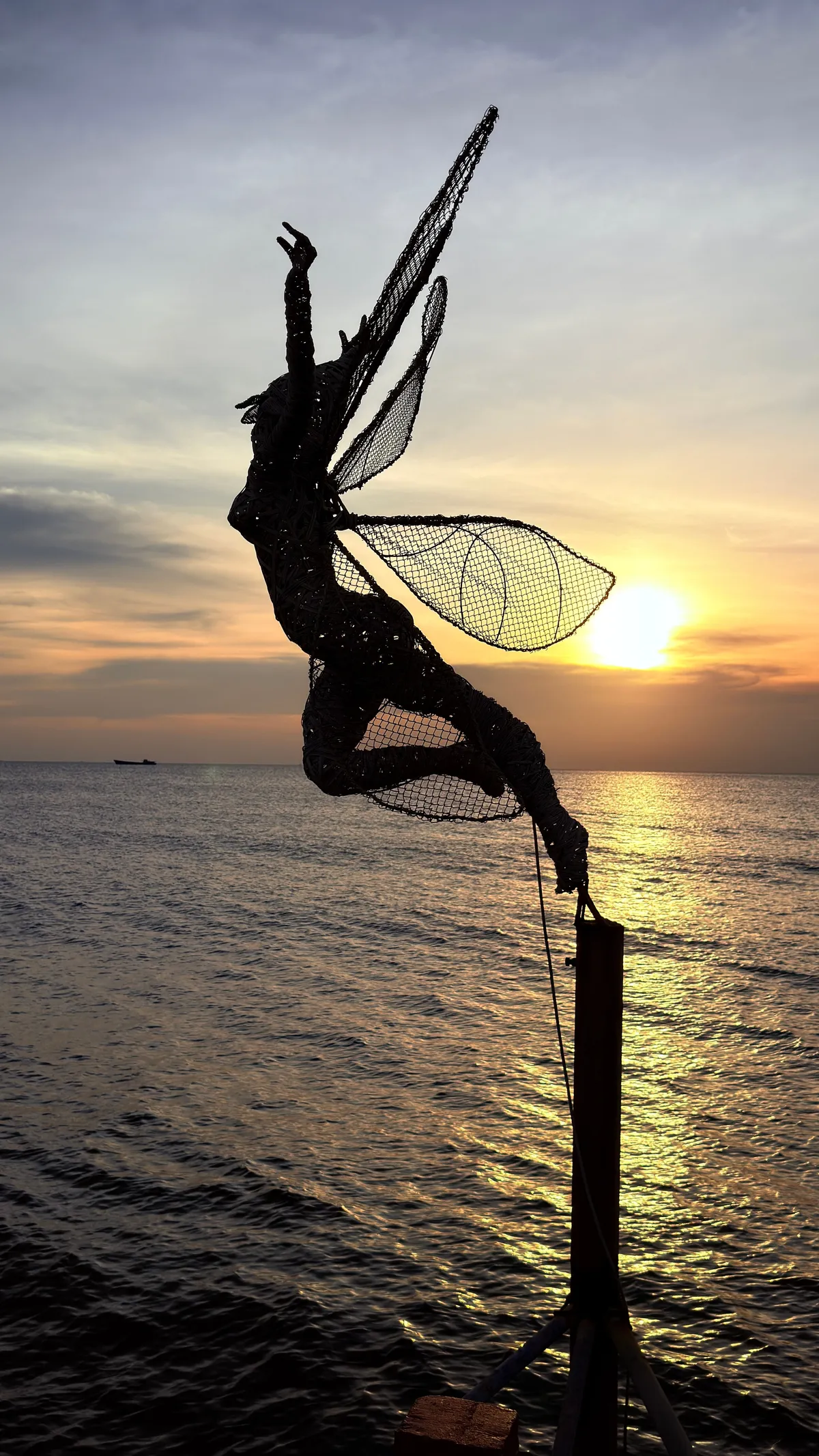 Wire sculpture of a fairy on a pole in silhouette against an orange sunset over the sea at Phu Quoc