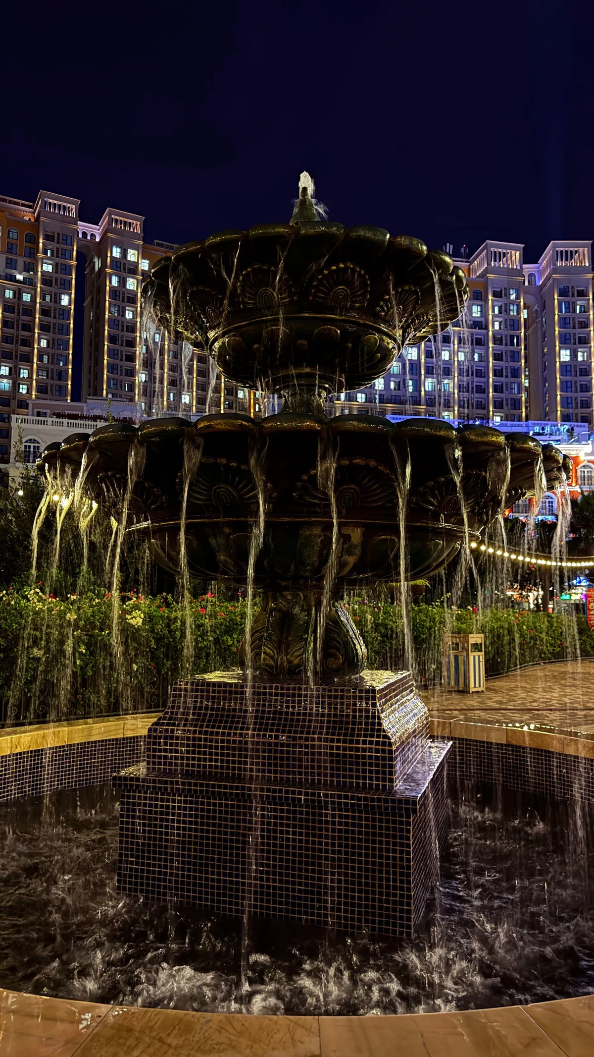 Tiered stone fountain at night with water pouring over its rim, hotel lights behind